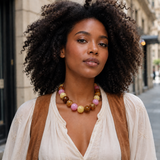 A woman with voluminous dark curly hair is shown from the chest up outdoors, wearing a chunky beaded necklace with pink, yellow, brown, and white beads, a cream blouse, and a brown vest. The background shows a softly blurred city street.