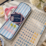 A mahjong set displayed on a marble tabletop, including a light blue and white striped carrying case with pink handles, a set of mahjong cards labeled 'Mahjong Cards 2026', and multiple tiles arranged on the table.