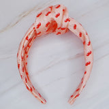 Soft satin headband adorned with a playful cherry and bow pattern, featuring vibrant red cherries and delicate bows on a pink background.