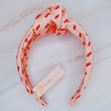 Soft satin headband adorned with a playful cherry and bow pattern, featuring vibrant red cherries and delicate bows on a pink background.