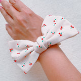 Playful hair scrunchie featuring a charming red cherry print and an attached bow tie, perfect for adding a sweet and fruity flair to ponytails or buns.
