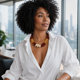 A woman with curly black hair wearing a white button-down shirt and a chunky beaded statement necklace with amber, brown, and cream beads, seated in an office with a city skyline in the background.