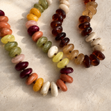 Three strands of polished gemstone beads in assorted colors, including amber brown, green, pink, orange, yellow, and cream, arranged on a textured beige surface.