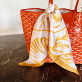 White scarf with gold patterns draped over a red handbag on a reflective surface.