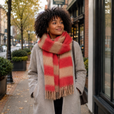 A woman with curly black hair stands on a city sidewalk wearing a thick, fuzzy striped scarf in red and beige, paired with a light gray coat.