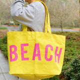 Person holding a yellow tote bag with 'BEACH' written in pink letters.