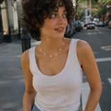 Woman standing on a city street wearing a white tank top and blue jeans.