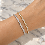 Two tennis-style bracelets on a person's wrist, one in silver with clear stones and one in yellow gold with clear stones, photographed over a white marble surface.