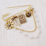 Gold-tone safety pin brooch adorned with floral and ribbon charms, delicate chain drapes, and pearl accents—offering a romantic, vintage-inspired accessory for bags, jackets, or scarves.
