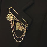 Gold-tone safety pin brooch adorned with floral and ribbon charms, delicate chain drapes, and pearl accents—offering a romantic, vintage-inspired accessory for bags, jackets, or scarves.