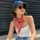 Woman wearing a blue cap, sunglasses, white tank top, denim shorts, and a red checkered scarf.