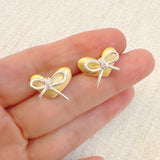 Delicate gold heart-shaped stud earrings adorned with a silver bow accent—perfect for adding a touch of charm and elegance to any look.