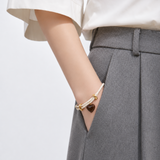 A close-up of a person's wrist wearing a double-strand cream cord bracelet with gold-toned hardware and a small dark brown heart charm, paired with a white top and gray trousers.