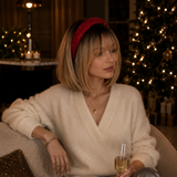 A woman sits on a couch in a cozy, warmly lit holiday setting, wearing a cream-colored fuzzy V-neck sweater, satin gray pants, and a red textured headband while holding a glass of champagne.