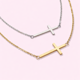 Two cross necklaces, one gold and one silver, on a light pink background.