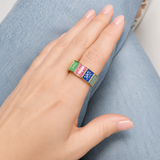 A close-up of a hand wearing a gold signet ring with three colorful enamel squares in green, pink, and blue, resting on light blue denim.