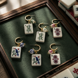 Six gold hoop earrings with rectangular mahjong tile charms and small bead drops, arranged on a dark green display tray. The charms feature white tiles with green bamboo, pink and blue bird motifs, and red circular symbols, with some matching earrings partially visible in a wooden case.