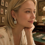 A blonde woman in a floral robe wearing a distinctive drop earring with a white enamel tile and green Chinese characters, posed in a warm, dimly lit room with mahjong tiles visible in the background.