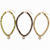 Three beaded necklace strands displayed side by side on a white background, each with a gold-tone clasp and a round clear stone pendant. The left necklace is olive green, the middle is dark brown, and the right is light tan.
