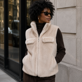 A woman wearing a cream faux-fur zip-front vest over a dark brown turtleneck, standing outdoors against a light stone building wall.