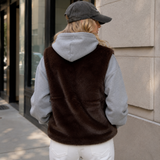 A woman viewed from behind wearing a dark brown faux-fur vest layered over a light gray hoodie, white pants, and a dark gray baseball cap, standing outdoors next to a stone building.