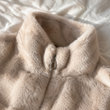 A close-up of a beige faux fur jacket with a high collar and a silver zipper pull, laid flat on a white textured fabric background.
