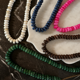 Five beaded necklaces in different colors arranged on a white scalloped dish over a dark marble surface: ivory, navy, bright pink, brown, and green. A small gold chain link is visible at the lower right.
