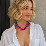 A woman with short blonde wavy hair wears a double-strand beaded necklace with pink and green beads, paired with an open white button-down shirt, posing against a concrete wall.