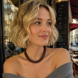 A blonde woman with short wavy hair wearing a dark brown beaded necklace and a gray off-the-shoulder cardigan, sitting at a cafe.