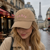 Woman wearing a beige cap with 'PARIS' in pink on a street with the Eiffel Tower in the background.