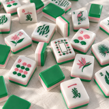 A close-up, top-down view of multiple Mahjong tiles scattered on a light fabric surface. The tiles are primarily white with green and pink layers, decorated with colorful Chinese characters, floral motifs, and symbols.