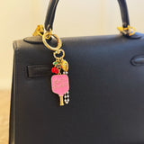 Playful bag charm featuring two pink pickleball paddles—one with cherries and one with “Let’s Pickle” text—accented with a cherry charm and gold-tone details for a fruity, sporty twist.