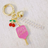 Playful bag charm featuring two pink pickleball paddles—one with cherries and one with “Let’s Pickle” text—accented with a cherry charm and gold-tone details for a fruity, sporty twist.