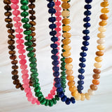 A collection of polished gemstone bead necklaces in various colors, including deep blue, green, brown, pink, and cream. Each necklace features smooth, rounded beads with a glossy finish, offering a timeless and versatile accessory look.