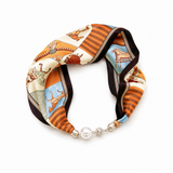 A silk scarf with a multicolor pattern of horses and stripes, styled in a loop with a silver-toned beaded chain and a white pearl clasp, on a white background.