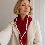 A woman wearing a cream-colored coat and a red knit scarf with a white scalloped edge, captured in a soft indoor setting.