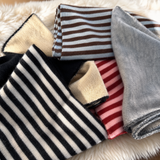 A pile of folded striped knit fabric scarves or wraps on a white faux-fur surface. Visible colorways include black and white stripes, brown and white stripes, solid light gray, red and white stripes, and a cream-colored knit with a black fuzzy trim.