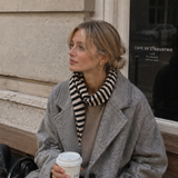 Woman in a gray coat with a striped scarf holding a coffee cup, sitting on steps in front of a building.