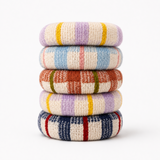 A stack of five knitted circular coasters or trivet mats in different multicolor stripe and plaid patterns, set against a plain white background.