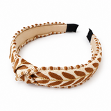 A wide fabric headband with a twisted knot front, featuring a brown and beige leaf pattern with gold glitter accents, a beige lining, and black adjustable end sliders, shown on a white background.