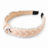A wide fabric headband with a knotted front detail, featuring a textured woven pattern in light peach and beige with light blue beaded trim, and black elastic end caps, shown on a white background.