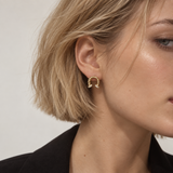A close-up of a model's ear wearing a small gold crescent-shaped hoop earring with tiny clear stones along the curve, against a plain light background.