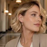 A close-up of a woman with blonde wavy hair wearing a small gold hoop earring, set against a softly blurred indoor hallway background.