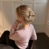 A woman with blonde hair styled in a low bun with a large cream satin scrunchie, wearing a light pink knit sweater and black pants, seated indoors.