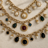 An antique-style gold chain necklace with multiple charms, including round and teardrop-shaped white pearls, square and round black and green gemstones, and central oval red and black cabochons, displayed on a neutral beige background.