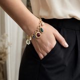 A gold charm bracelet with multiple colorful gemstone charms and pearl accents, shown worn on a woman's wrist with her hand in her pocket.