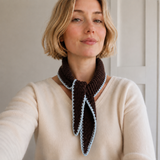 A woman wearing a cream-colored knit sweater and a dark brown knitted scarf with a light blue edge, posing in a neutral indoor setting.