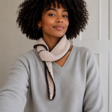 A woman with curly black hair wearing a light gray V-neck sweater and a cream-colored knit scarf with dark brown trim, posing indoors against a white paneled wall.