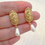 Gold oval stud earrings featuring a detailed cross design and dangling teardrop pearls—blending vintage charm with timeless elegance.