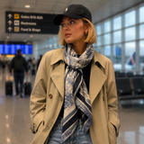 A woman wearing a beige trench coat, a blue and white patterned scarf, a black baseball cap, and blue jeans stands in an airport terminal.